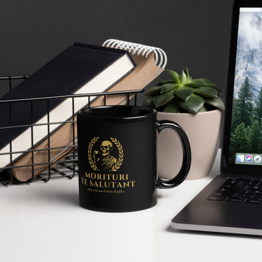 Tasse MORITURI TE SALUTANT – 11 oz im Büro-Setting – goldener Totenkopf im Lorbeerkranz auf schwarzer Glanzkeramik, powered by Nyrra-Design