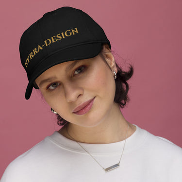Frau trägt schwarze Bio-Cap mit goldener Nyrra-Design-Stickerei, Frontansicht vor rosa Hintergrund.